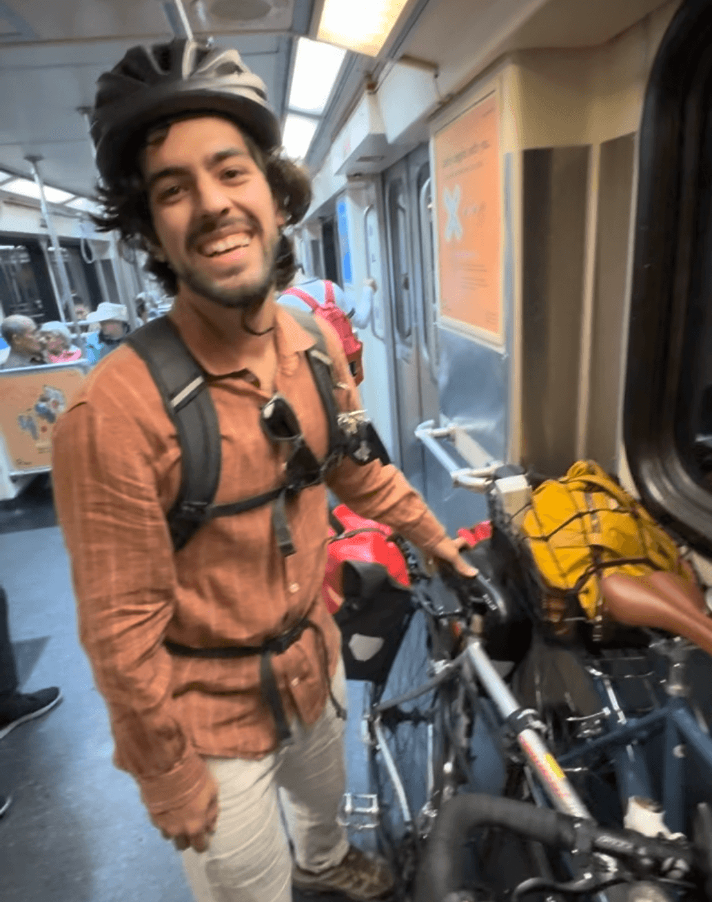 Nejat Can, wearing a bike helmet, backpack, and rust-colored shirt, smiling aboard a train car while holding a touring bike loaded with red and yellow panniers. Other passengers are seated in the background.